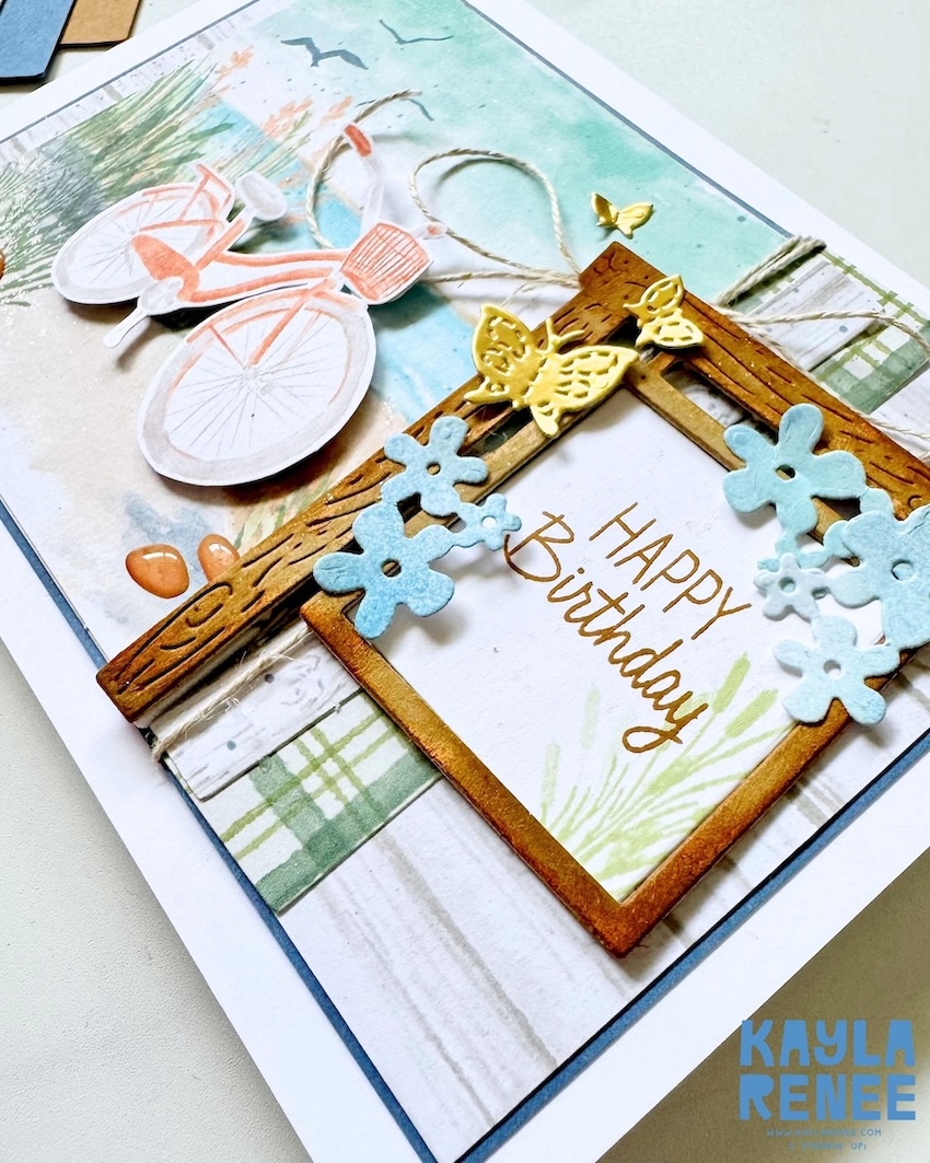 Angled close-up of a handmade birthday card made with the Stampin’ Up! Lakeside Retreat Paper Pumpkin Kit, showcasing layered woodgrain frames, soft blue flowers, gold butterfly embellishments, twine details, and a lakeside bicycle scene.
