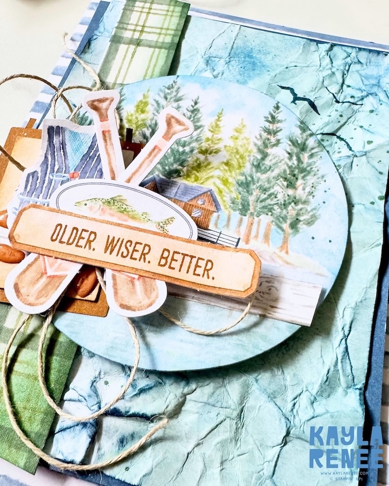 Close-up of a rustic handmade card made with the Stampin’ Up! Lakeside Retreat Paper Pumpkin Kit, featuring fishing-themed embellishments, cabin imagery, layered circles, natural twine, and textured watercolour-style backgrounds.