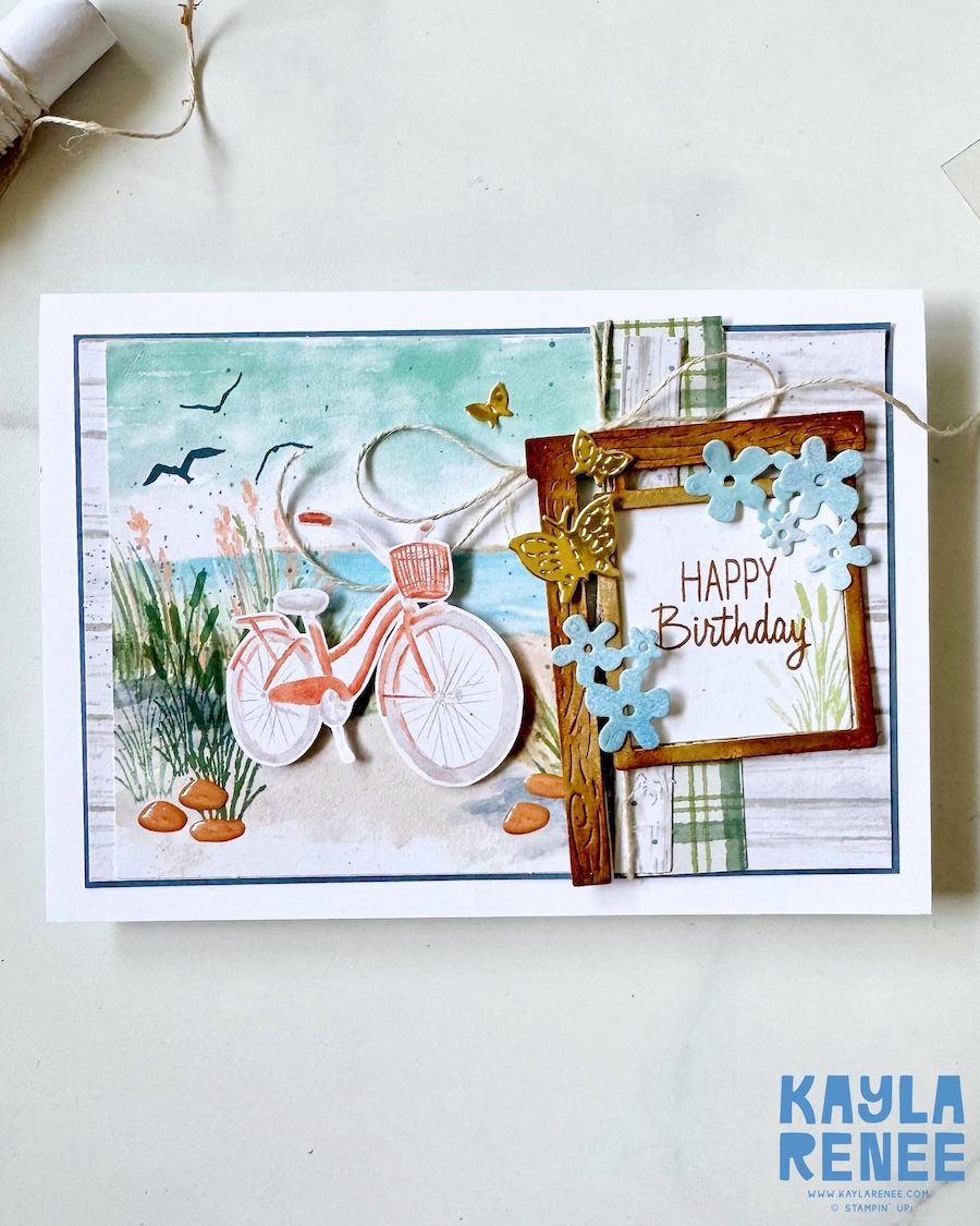 Handmade birthday card created as an alternative project using the Stampin’ Up! Lakeside Retreat Paper Pumpkin Kit, featuring a pastel lakeside scene with a bicycle, rustic wood frame, gold butterflies, layered florals, and a “Happy Birthday” sentiment.