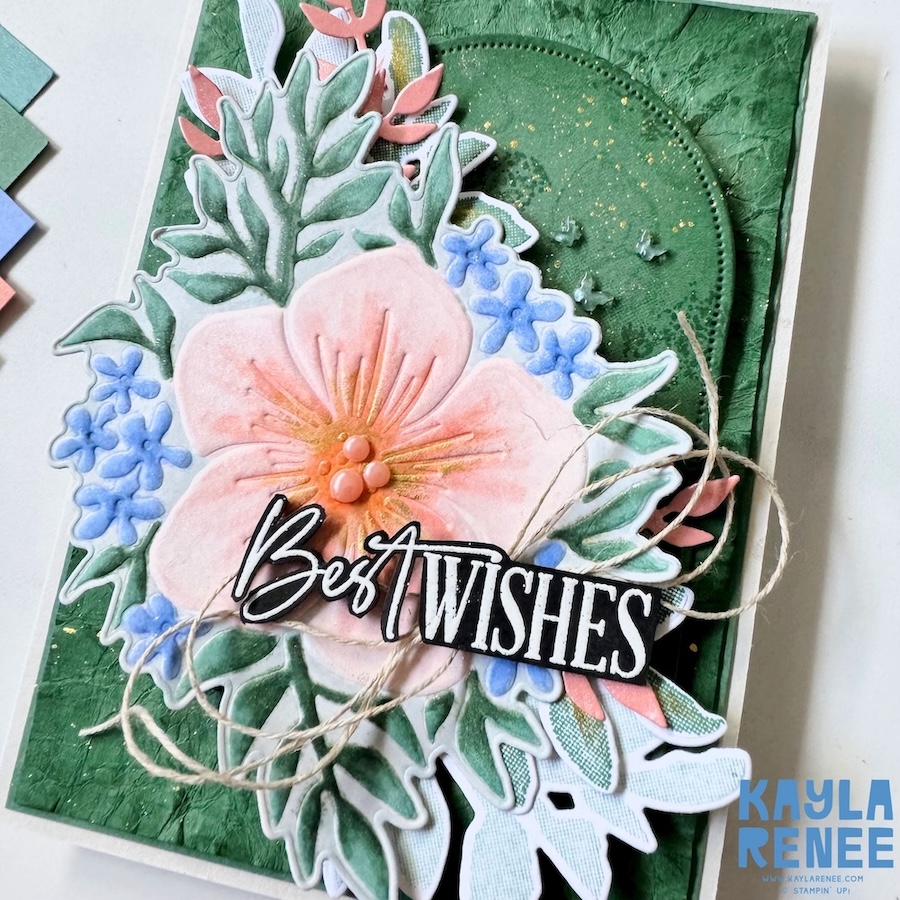 Angled close-up of a Stampin’ Up! Opulent Oasis bundle card featuring dimensional floral layers, blended green foliage, delicate blue flowers, rustic twine loops, and a black-and-white “Best Wishes” sentiment.