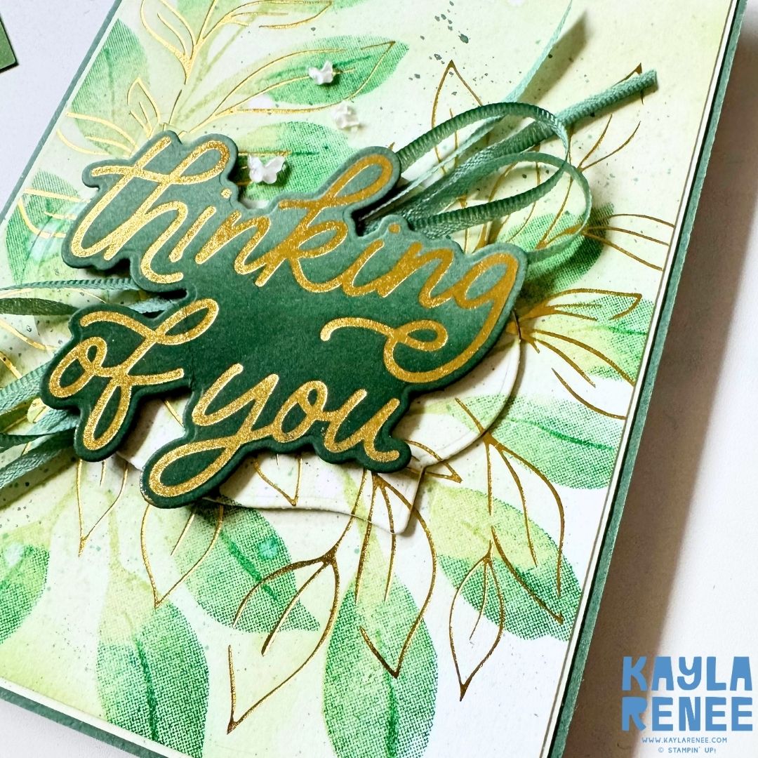 Close-up of a handmade Stampin’ Up! Botanical Glow hot foil card featuring shimmering gold foiled leaves and a bold Classic Greetings gold foiled "Thinking of You" sentiment. Soft green ink blending, layered ribbon, subtle ink splatter, and dimensional embellishments highlight the luxurious hot foiling techniques and botanical styling.