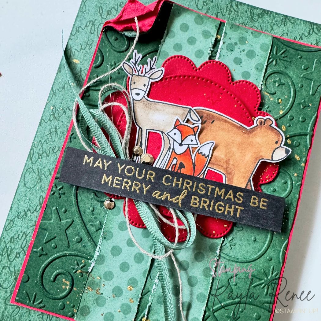 Angled view of a handmade Christmas card showcasing layered textures created with the Stampin’ Up! Woodland Wishes stamp set and Swirls of Magic Embossing Folder. The rustic design features embossed green cardstock, torn patterned paper, hand-coloured woodland animals, stitched die-cut circles, ribbon, linen thread, metallic embellishments and a gold heat-embossed Christmas sentiment.