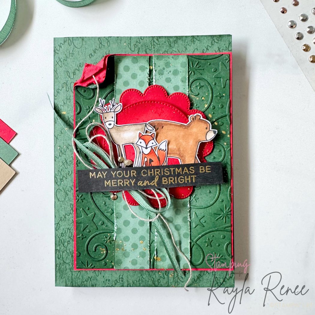 Handmade Christmas card created for The Stamping Lounge Christmas in July event featuring the Stampin’ Up! Woodland Wishes stamp set and Swirls of Magic Embossing Folder. The design combines embossed and distressed green panels with festive red layers, woodland animals, gold splatter, ribbon, linen thread and a gold heat-embossed "May Your Christmas Be Merry and Bright" greeting for a rustic Christmas look.