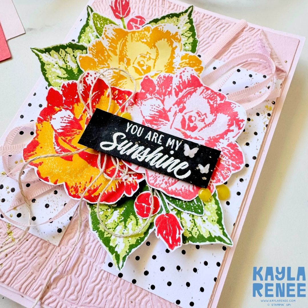 Close-up of a colourful Stampin’ Up! Begonia Belle floral card featuring layered yellow, orange, red and pink flowers surrounded by green leaves. A black “You are my Sunshine” sentiment is layered across the floral arrangement with pink ribbon, twine and small embellishments.