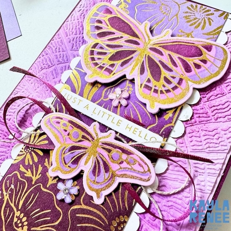 Angled close-up of a handmade Stampin’ Up! hot foil card in shades of purple, burgundy, white and gold. The design features two dimensional butterflies created with the Butterfly Flight hot foil dies, layered over gold-foiled floral imagery from the Garden Silhouette hot foil dies. Textured cardstock, scalloped edging, ribbon, linen thread and small floral embellishments create a richly layered design around the “Just a Little Hello” sentiment.
