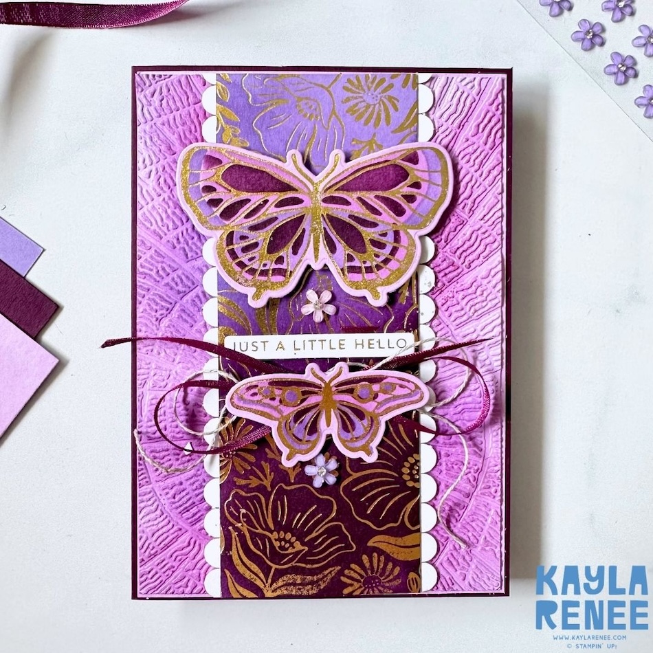 Handmade purple and gold greeting card featuring the Stampin’ Up! Garden Silhouette and Butterfly Flight hot foil dies used with the Stampin’ Up! Hot Foil System. The card has a textured purple background, a central strip of purple and gold foiled floral paper framed with white scalloped edges, and two dimensional butterflies with intricate gold hot foil details. Burgundy ribbon, linen thread and small purple flowers add texture and dimension, with a “Just a Little Hello” sentiment across the centre.