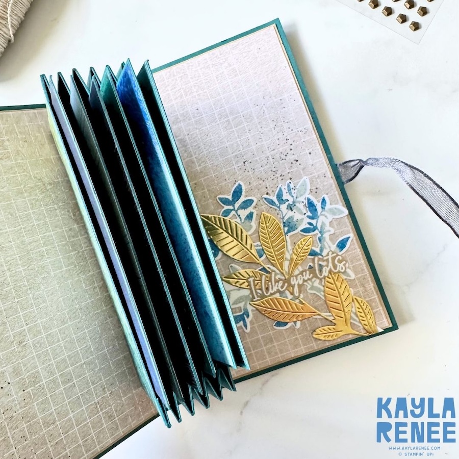 Pop-up expanding pocket card opened to reveal the accordion-style central structure, patterned paper panels and a decorated writing panel featuring layered blue and gold botanical elements.