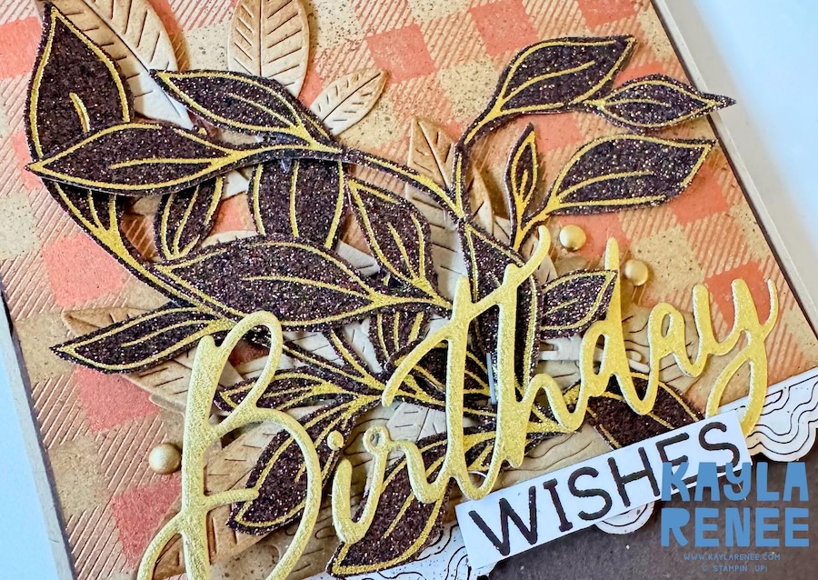 Close-up of a handmade birthday card featuring a neutral colour palette, layered dark foliage with gold hot foil accents, natural-toned leaves, textured patterned paper and a large gold “Birthday” sentiment with “Wishes” underneath.