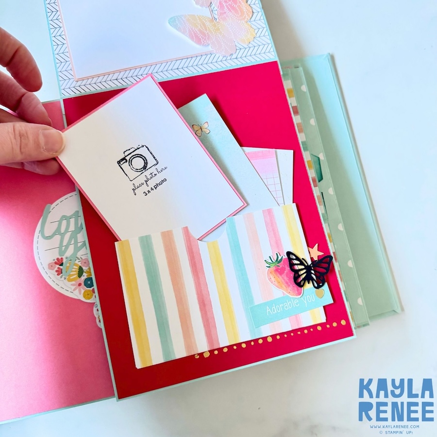 Inside the handmade mini album showing a bright pink scrapbook page with layered photo mats, pastel striped paper, butterfly embellishments and a small “Adorable you” sentiment.