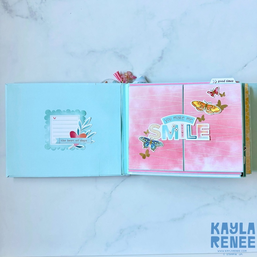Open view of the handmade mini album showing a light aqua interior cover beside a pink scrapbook page decorated with butterflies and the phrase “you make me SMILE.”