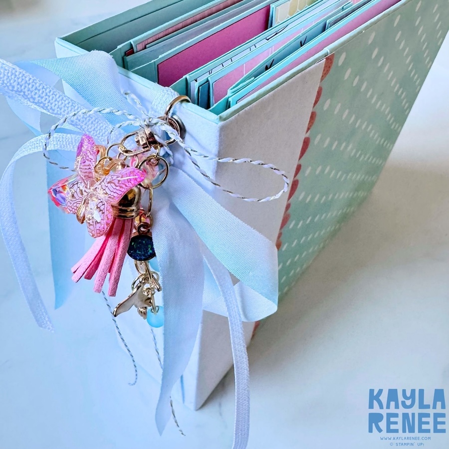 Side view of the handmade Stampin’ Up! mini album showing its layered pages, light aqua cover, white ribbon binding and decorative charms including a pink butterfly, tassels and beads.