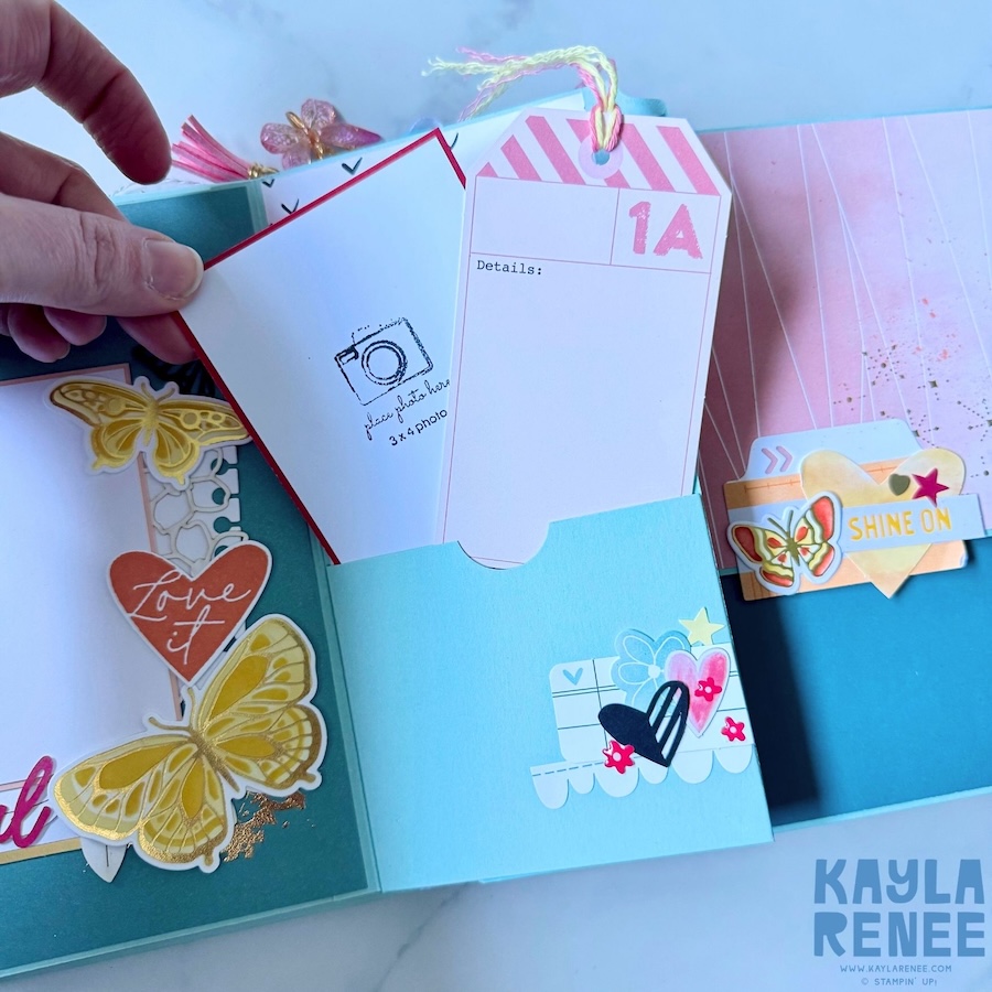 Open mini album page featuring a large pink and blue watercolour background, a lined journaling card, layered butterfly embellishments and a pocket containing additional photo and journaling pieces.