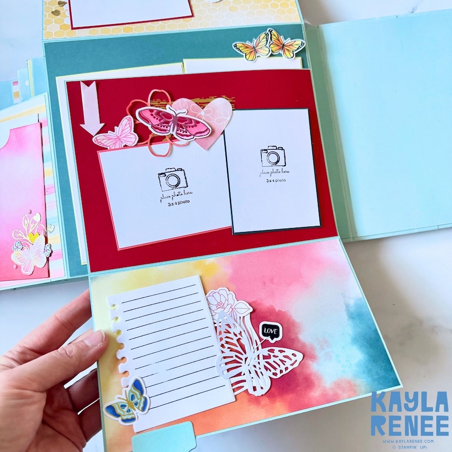 Open mini album page featuring a large pink and blue watercolour background, a lined journaling card, layered butterfly embellishments and a pocket containing additional photo and journaling pieces.